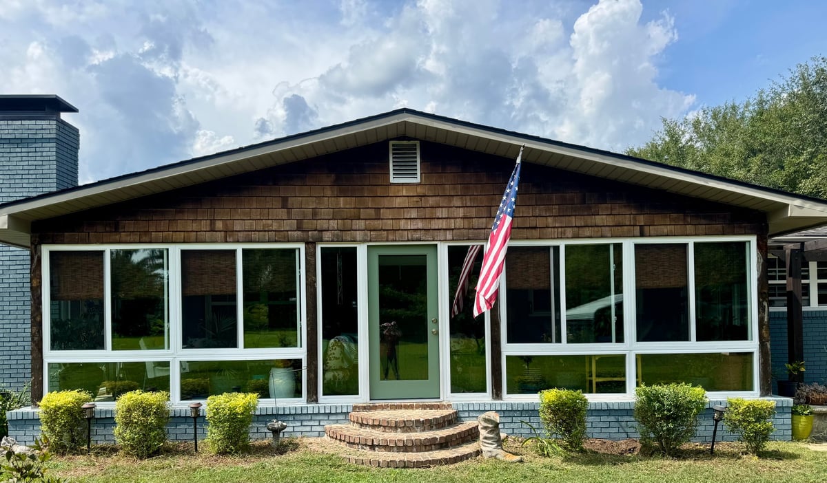 Modern single-story house with large windows, brick foundation, American flag, and landscaped shrubs in front