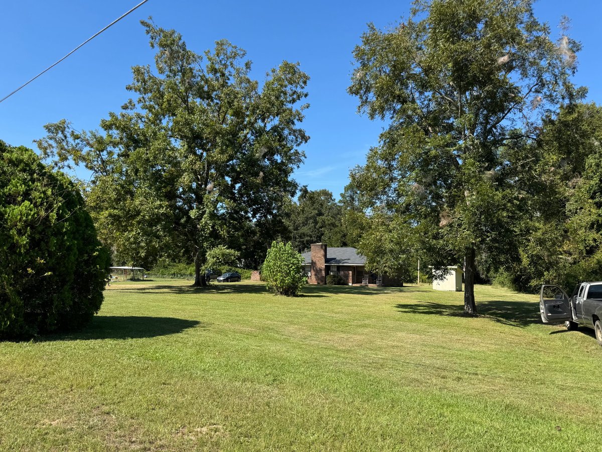 Rural property with brick house, large green lawn, mature trees, and clear blue sky