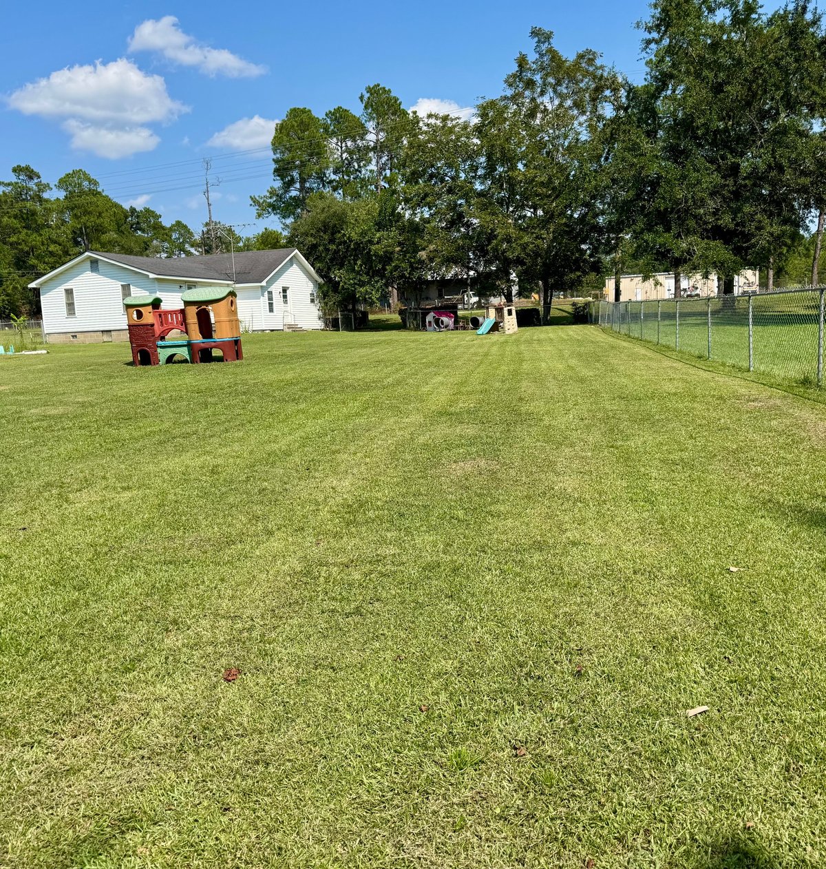 Rural farm property with white buildings, red barn, green grass field, wooden playground, and trees under blue sky