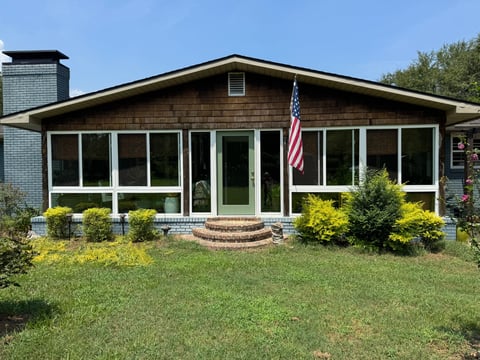Front view of a single-story brown brick house with white trim windows, American flag, brick steps, and landscaped yard with green shrubs.