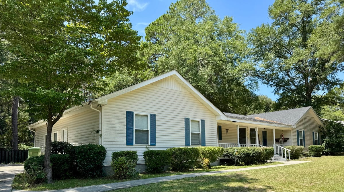 Single-story beige house with blue shutters, front porch, and mature trees on a sunny day